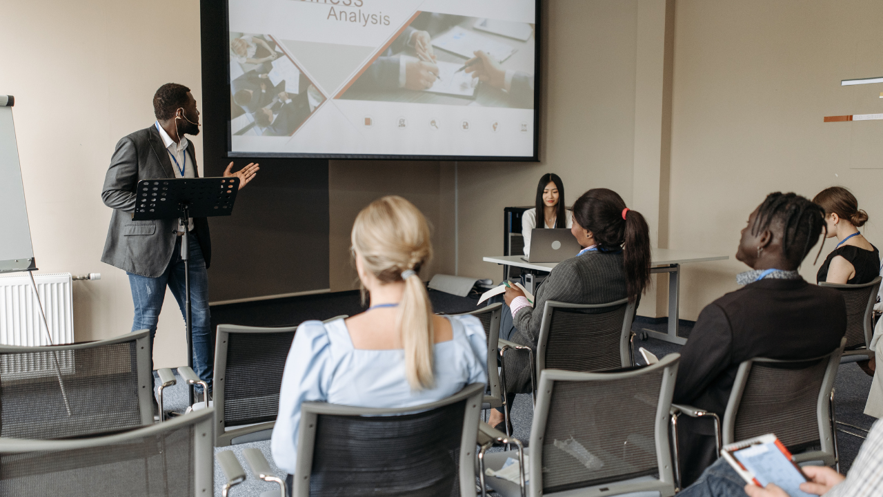 A speaker presenting a business analysis to a diverse group of professionals in a conference room, with a projected slide behind him and attendees seated and engaged.