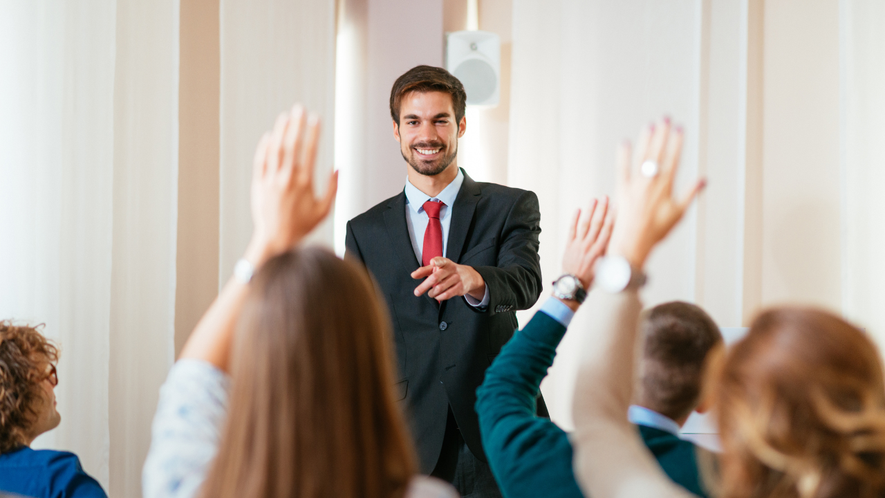 Man in a suit leading a seminar, smiling and pointing as attendees raise their hands.