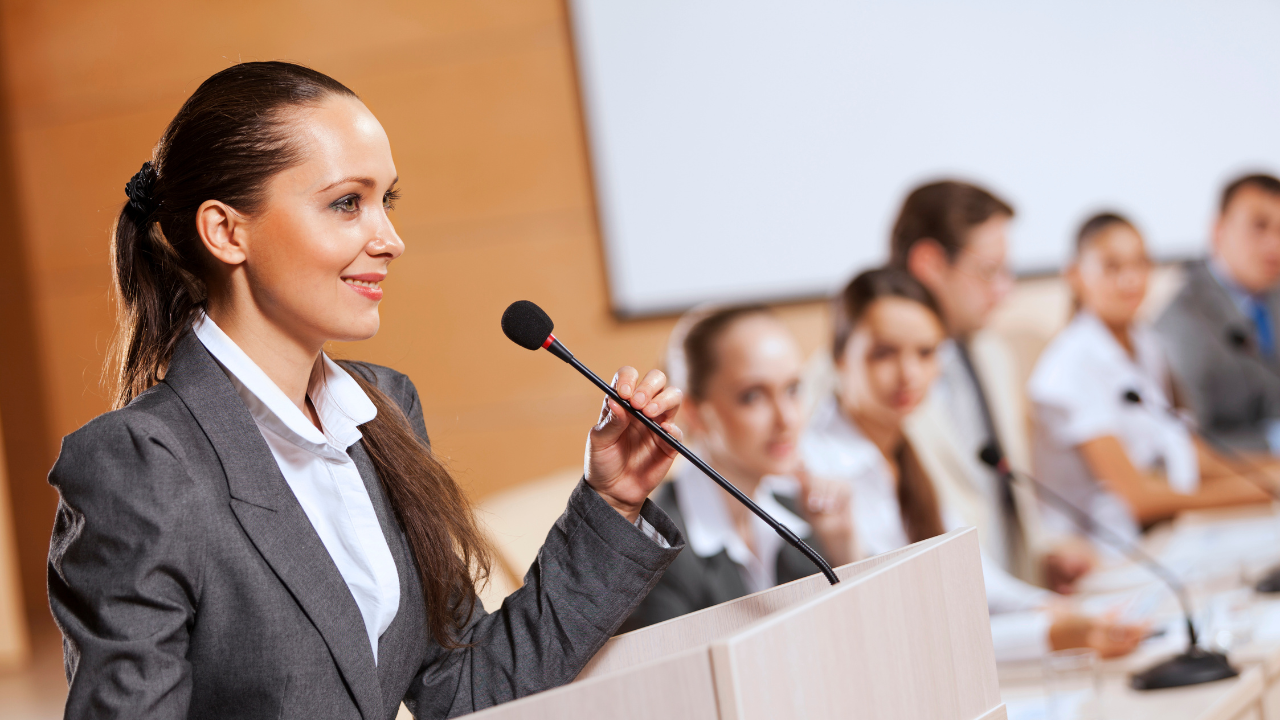 A confident woman in a suit speaking at a podium with a microphone, while a panel of professionals listens in the background.