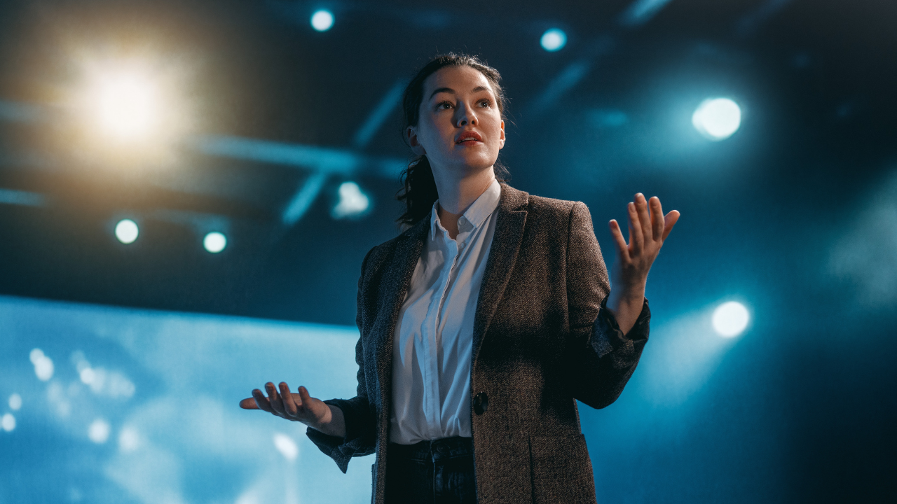 A confident woman with dark hair speaking on stage under blue spotlights, gesturing with open hands to an audience.