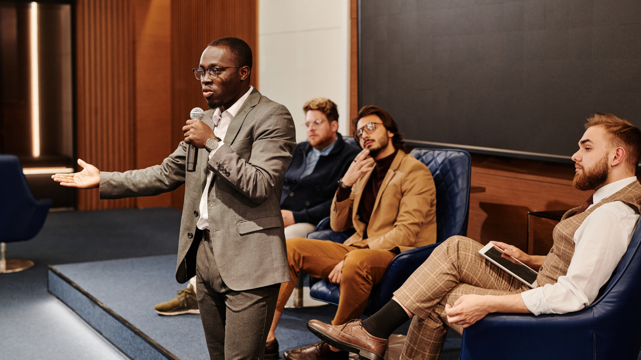 A man in a suit speaks into a microphone on a stage, gesturing with his left hand, while an audience listens in chairs to his right. Gaby Mammone.