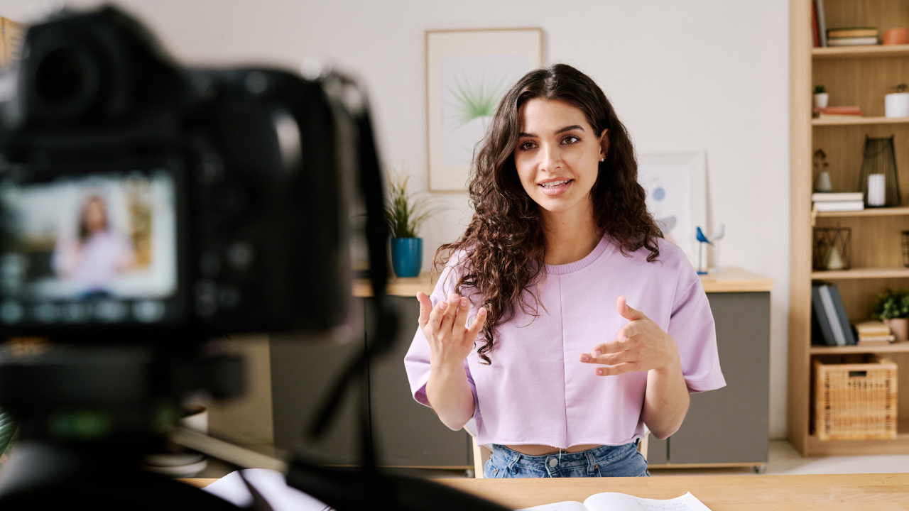 A young woman with curly hair is recording a video, speaking and gesturing in front of a camera on a tripod, in a home office setting. Gaby Mammone, speaking coach.