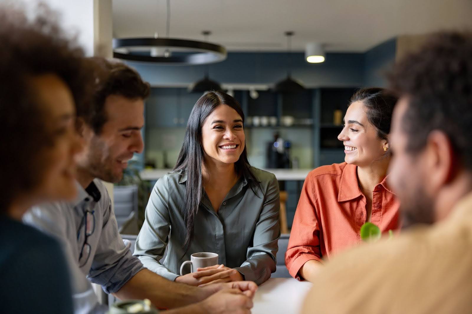 A diverse group of professionals smiling and engaged in conversation around a table in a modern office or meeting space. Gaby Mammone.