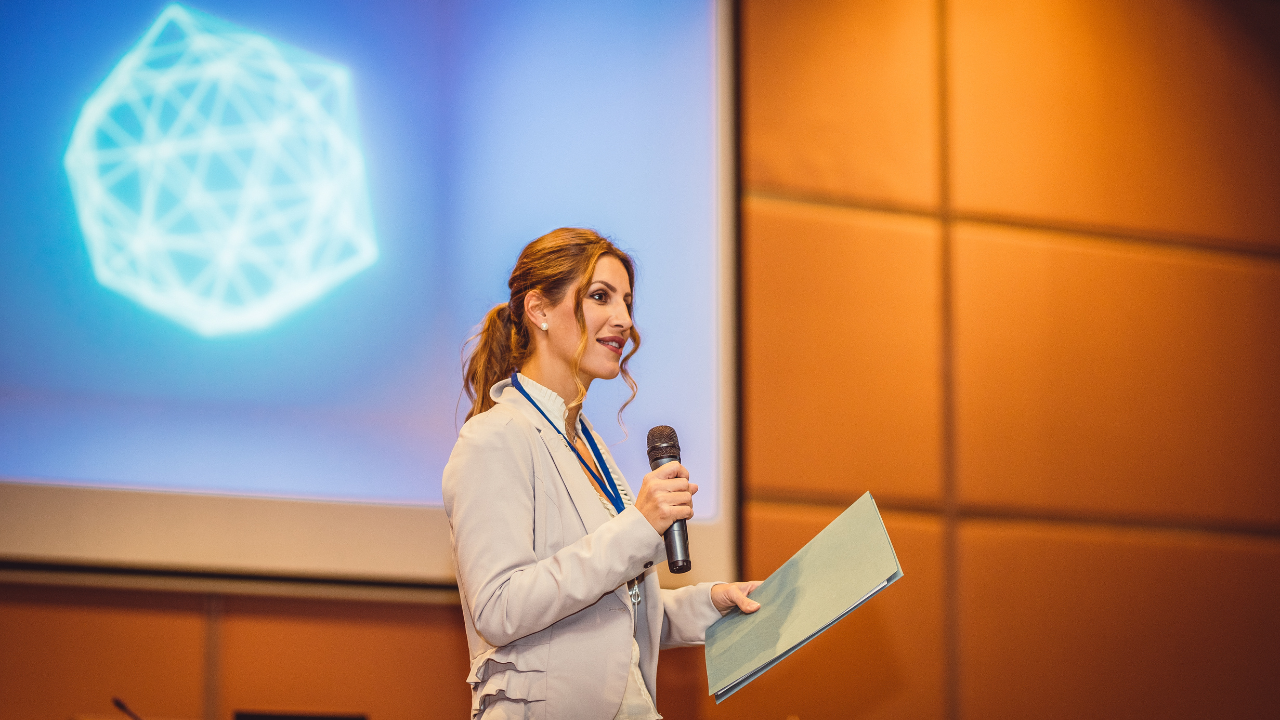 A woman presents with a microphone on a stage, in front of a screen, a skill Gaby Mammone is well known for.