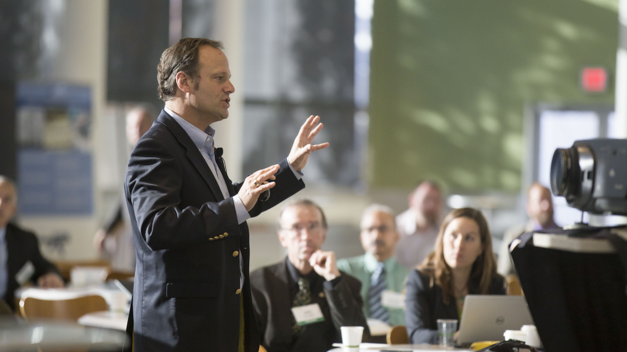 A man in a dark suit speaks passionately to an audience, demonstrating techniques similar to the ones Gaby Mammone teaches for confident public speaking